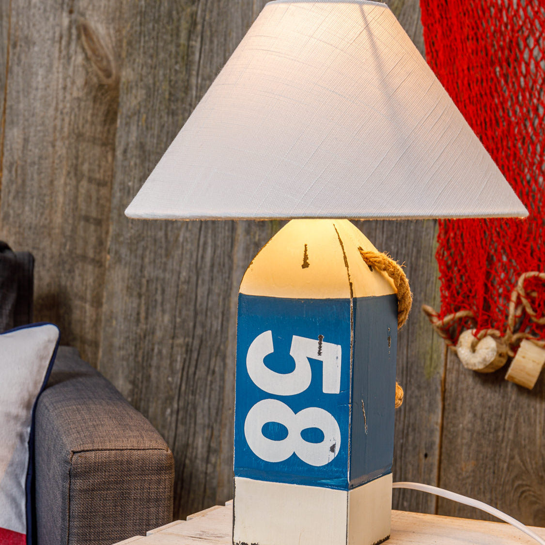 Tischlampe mit maritimem Design, blauem Sockel und Nummer 58, auf einem Holztisch vor Holzwand und roten Netz.