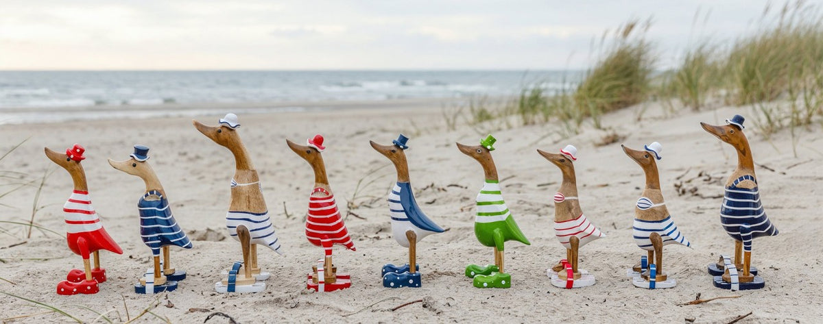Holzenten in gestreifter Badebekleidung stehen in einer Reihe am Strand mit dem Meer im Hintergrund.
