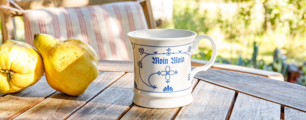Weiße Tasse mit blauem Muster und "Moin Moin" Aufschrift auf einem Tisch neben zwei gelben Quitten im Freien.